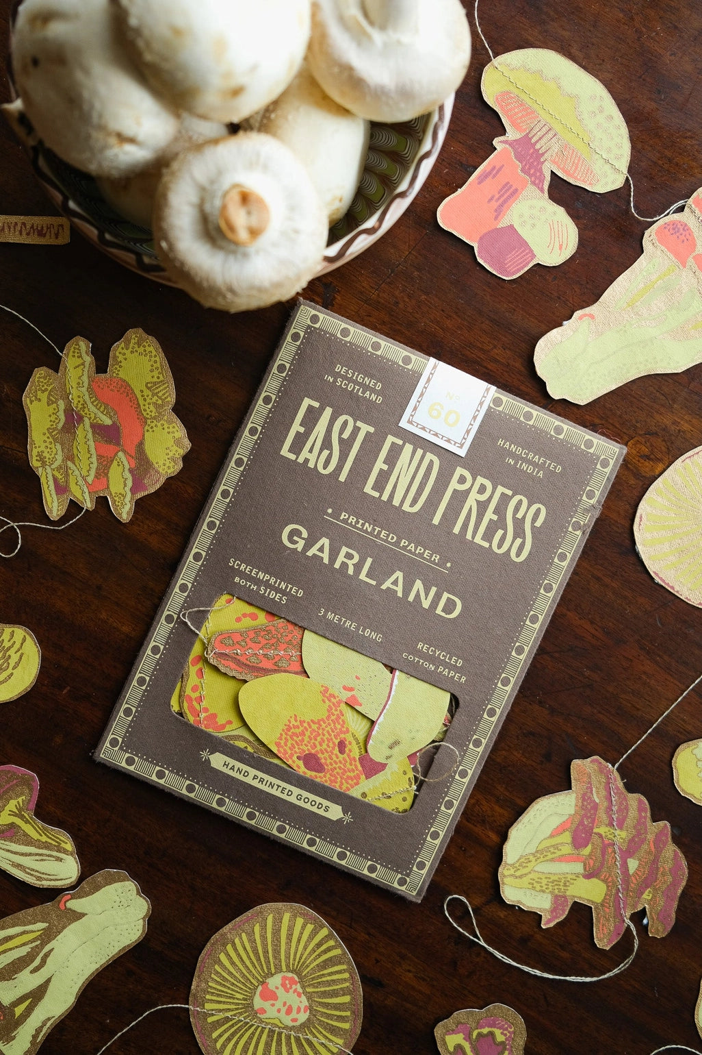 East End Press Garland package placed on a dark wooden surface, surrounded by mushroom-shaped paper garland pieces and a basket of white mushrooms. The package reads 'East End Press Garland' and shows illustrated mushroom designs on the front.