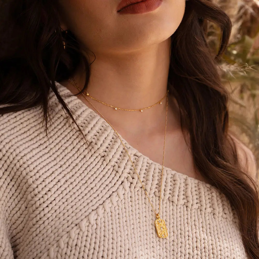 Close-up of a person wearing a gold necklace with a pendant, set against a blurred natural background.
