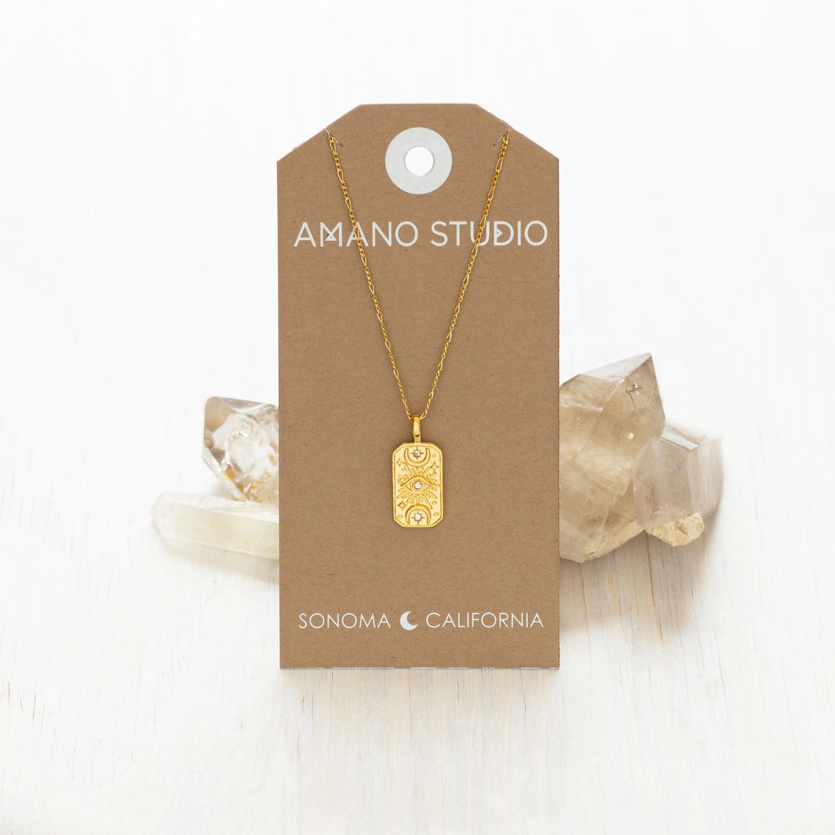 A gold rectangular pendant necklace with an engraved floral pattern, hanging from a brown kraft display card labeled 'AMANO STUDIO' and 'SONOMA CALIFORNIA'. The card is styled on a white wooden surface with clear crystal clusters behind it.