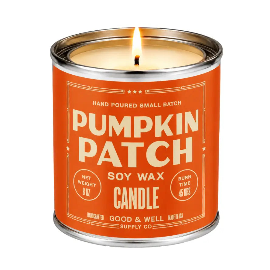 A lit 8 oz soy wax candle in a metal tin canister with a bright orange label reading 'Pumpkin Patch' by Good & Well Supply Co.
