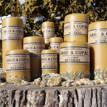 A collection of 'Sage & Copal' beeswax candles by Ugly Clover Candles, displayed on a rustic tree stump with natural elements and snow-dusted greenery in the background.