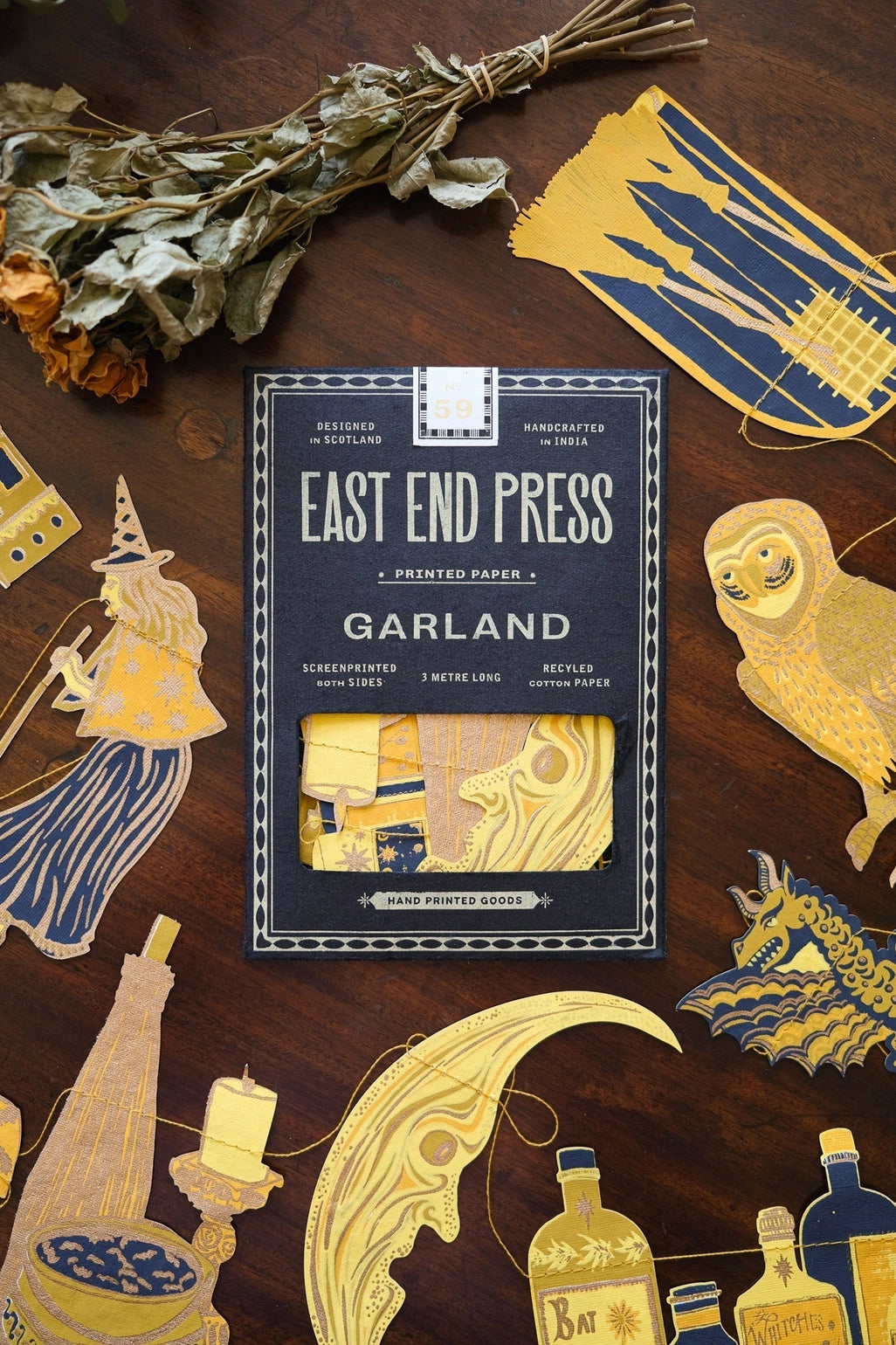 A styled flat-lay of the East End Press Garland printed paper product in the center, surrounded by decorative illustrated paper cutouts and dried flowers on a dark wood surface.