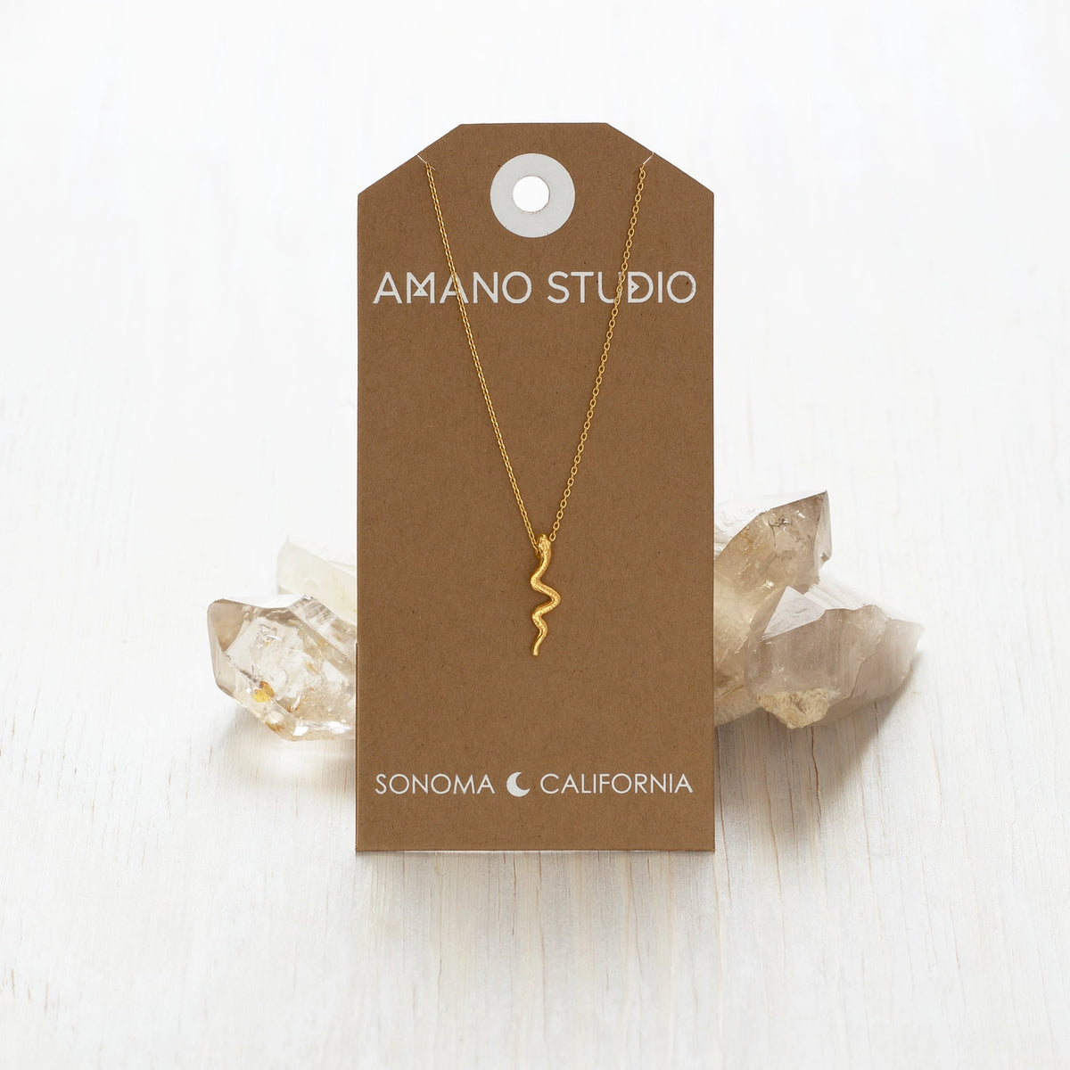 Gold necklace on a 'Amano Studio' branded card with crystals in the background