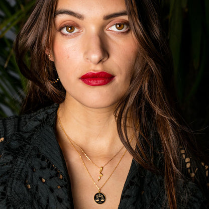 A close-up portrait of a woman with long dark brown hair, wearing layered gold necklaces, one with a black scale of justice pendant, and red lipstick, set against a dark background with green foliage.