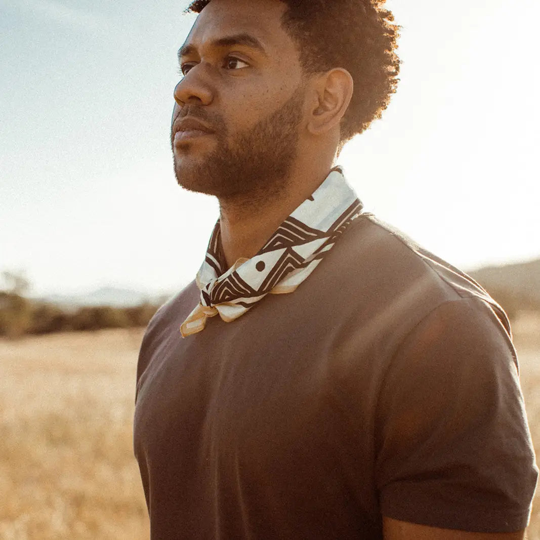 A man in a brown t-shirt wearing a white and brown geometric patterned neck scarf, photographed outdoors in warm sunlight.