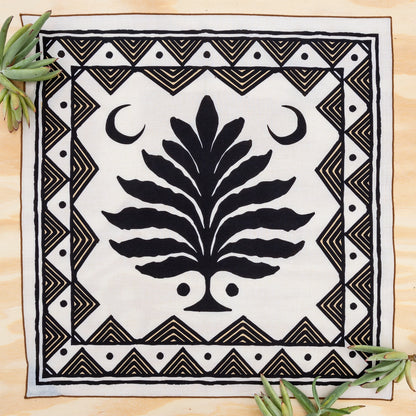 A square white textile with a bold black central tree motif, crescent moons, and geometric border patterns, laid flat on a light wood surface with small green leaves in the corners.