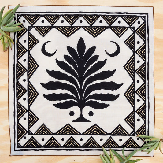 A square white textile with a bold black central tree motif, crescent moons, and geometric border patterns, laid flat on a light wood surface with small green leaves in the corners.